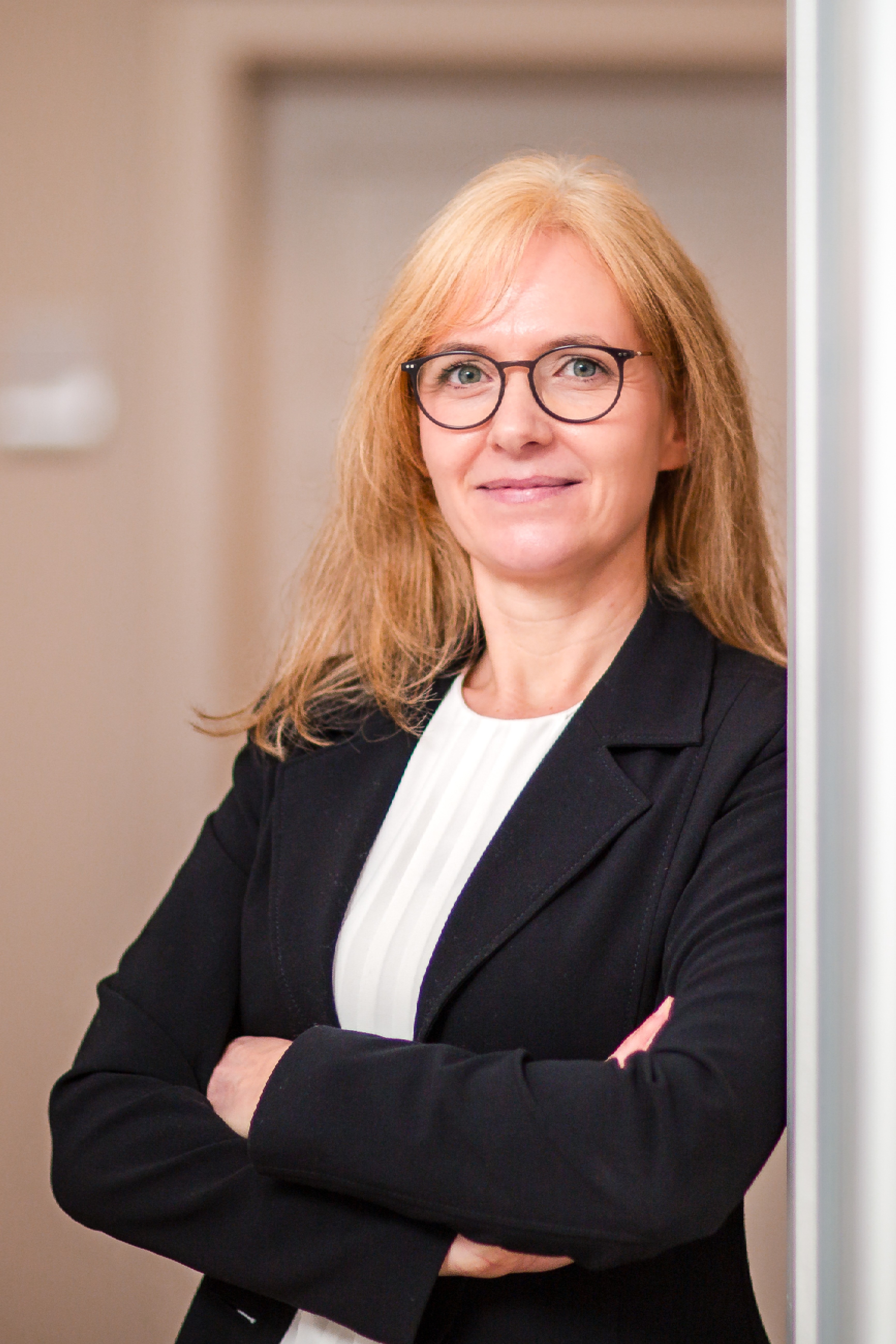 Eine Frau mit langen blonden Haaren und Brille lehnt lächelnd an einer Wand. Sie trägt ein schwarzes Jackett über einem weißen Oberteil und hat die Arme verschränkt.